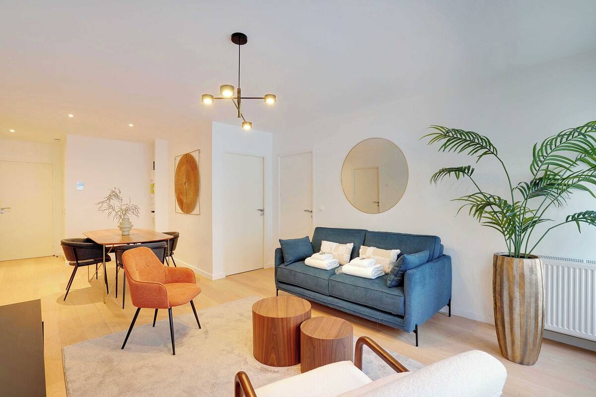 Una sala de estar moderna con un sofá azul, un sillón naranja, una mesa de centro de madera y una planta grande en maceta.