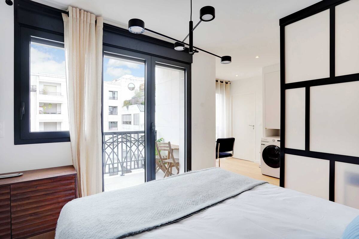 Un dormitorio moderno con una cama grande, un aparador de madera, un balcón con vistas a edificios y una lavadora en una esquina.