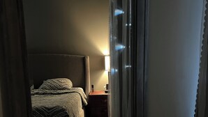En smal hall leder in till ett sovrum med en säng, ett nattduksbord och en lampa.