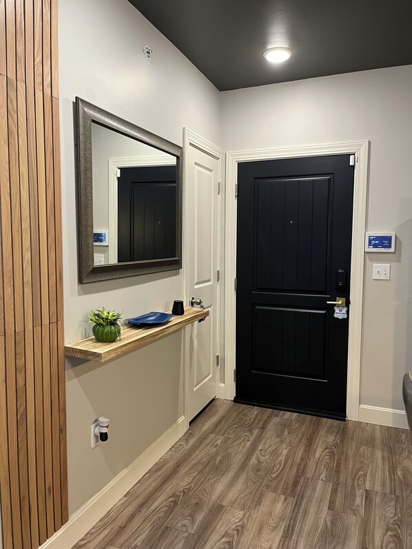 A hallway with a wooden panel wall, a mirror, a shelf with a plant and a mug, a black door with a security lock, and a thermostat on the wall.