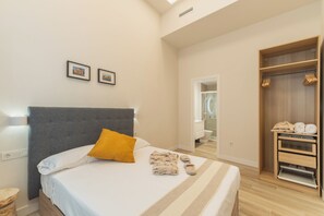 Ein ordentlich bezogenes Bett mit grauem Kopfteil, einem gelben Kissen und einem Paar Hausschuhen darauf. Über dem Bett hängen zwei gerahmte Bilder an der Wand.