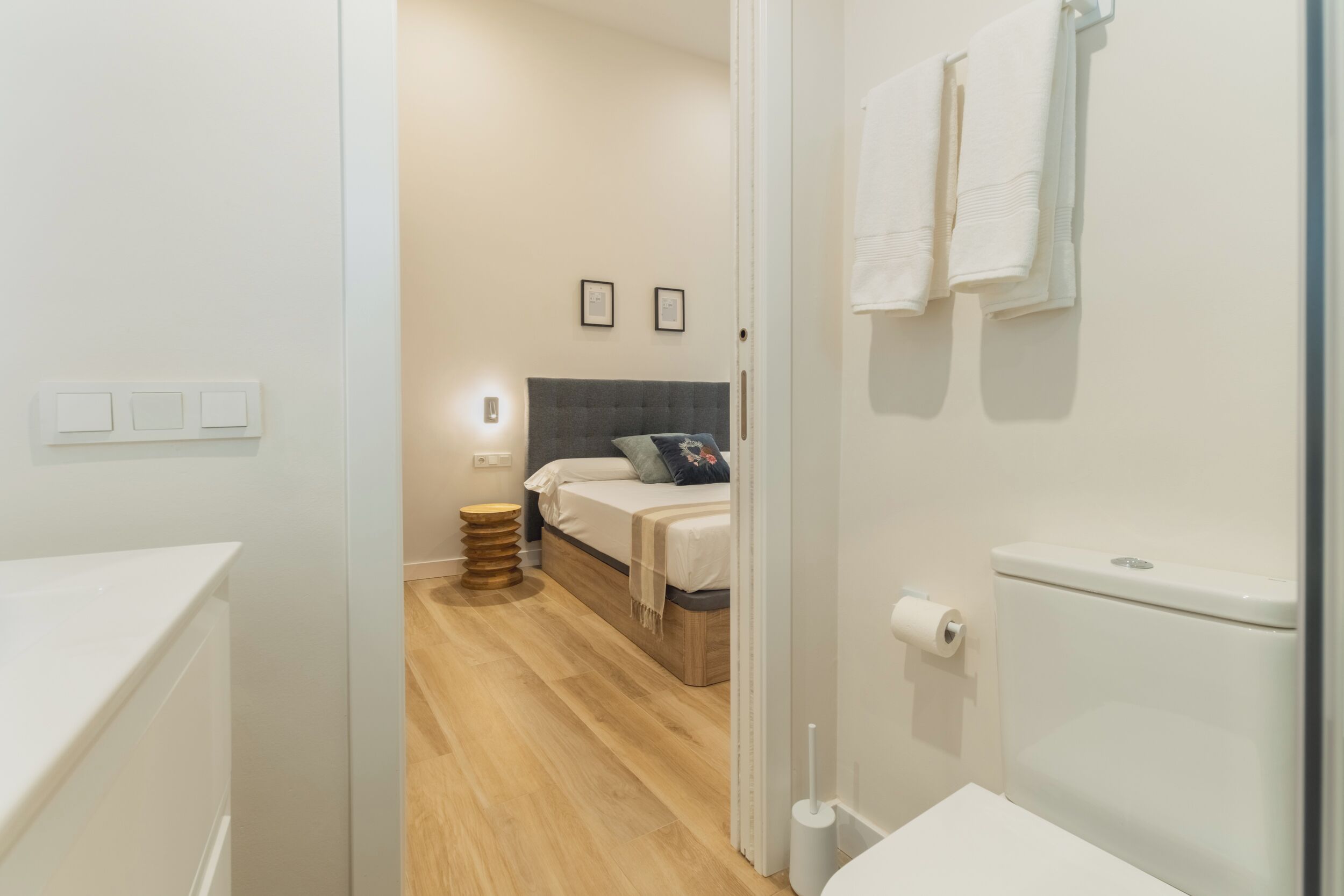 A modern hotel room with a wooden floor, a bed with a grey headboard, a bedside table, and a white toilet.