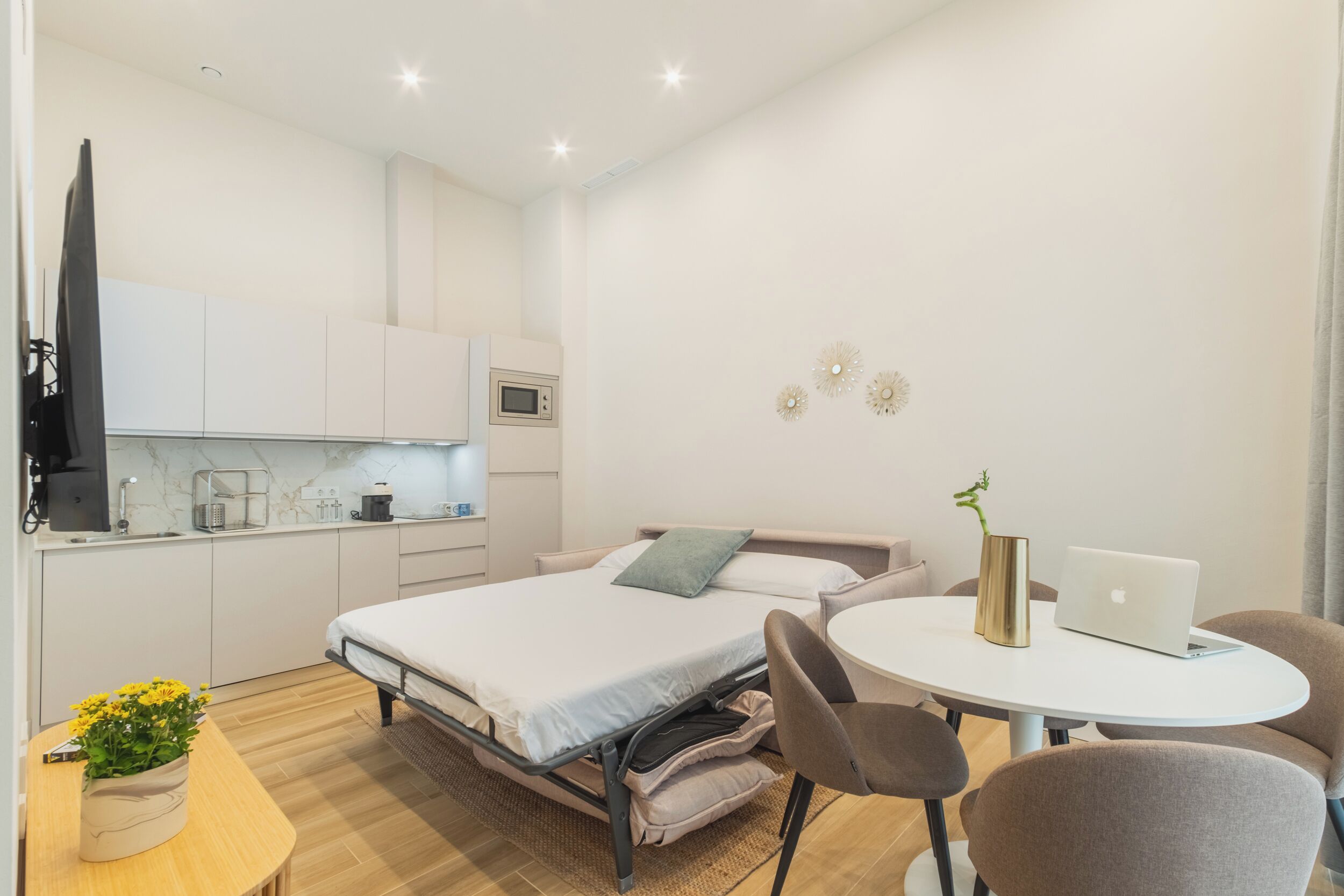 A modern, compact hotel room with a bed, bedside table, laptop, and a small round table with chairs.