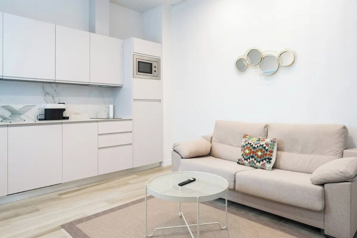Una sala de estar moderna con un sofá, una mesita redonda y una cocina con alacenas blancas y un microondas.
