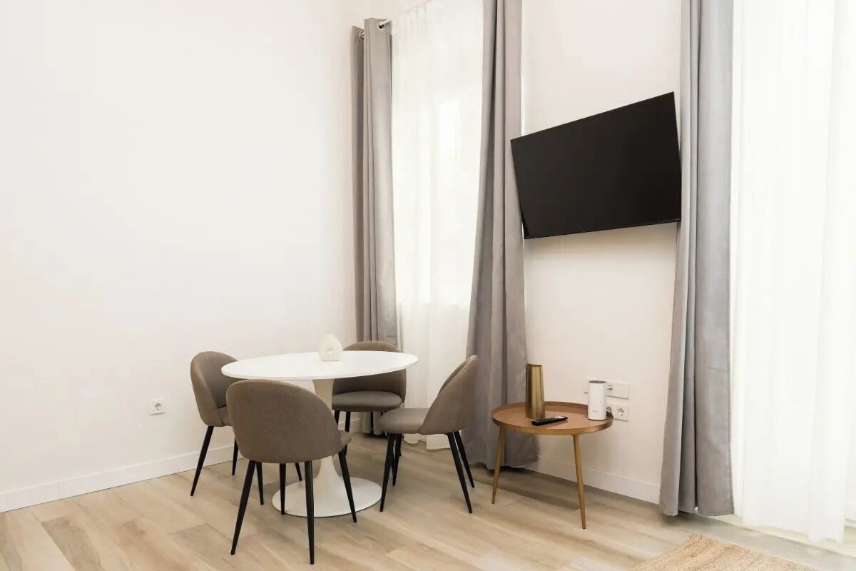 A modern room with a round dining table, four chairs, a small side table, a flat-screen TV mounted on the wall, and light-colored curtains.