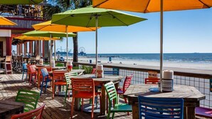 A beachside restaurant with colorful chairs and umbrellas, wooden deck, and ocean view. - SPEND YOUR SPRING BREAK IN A HOT TUB/HEATED POOL on the ISLAND! (Galveston)