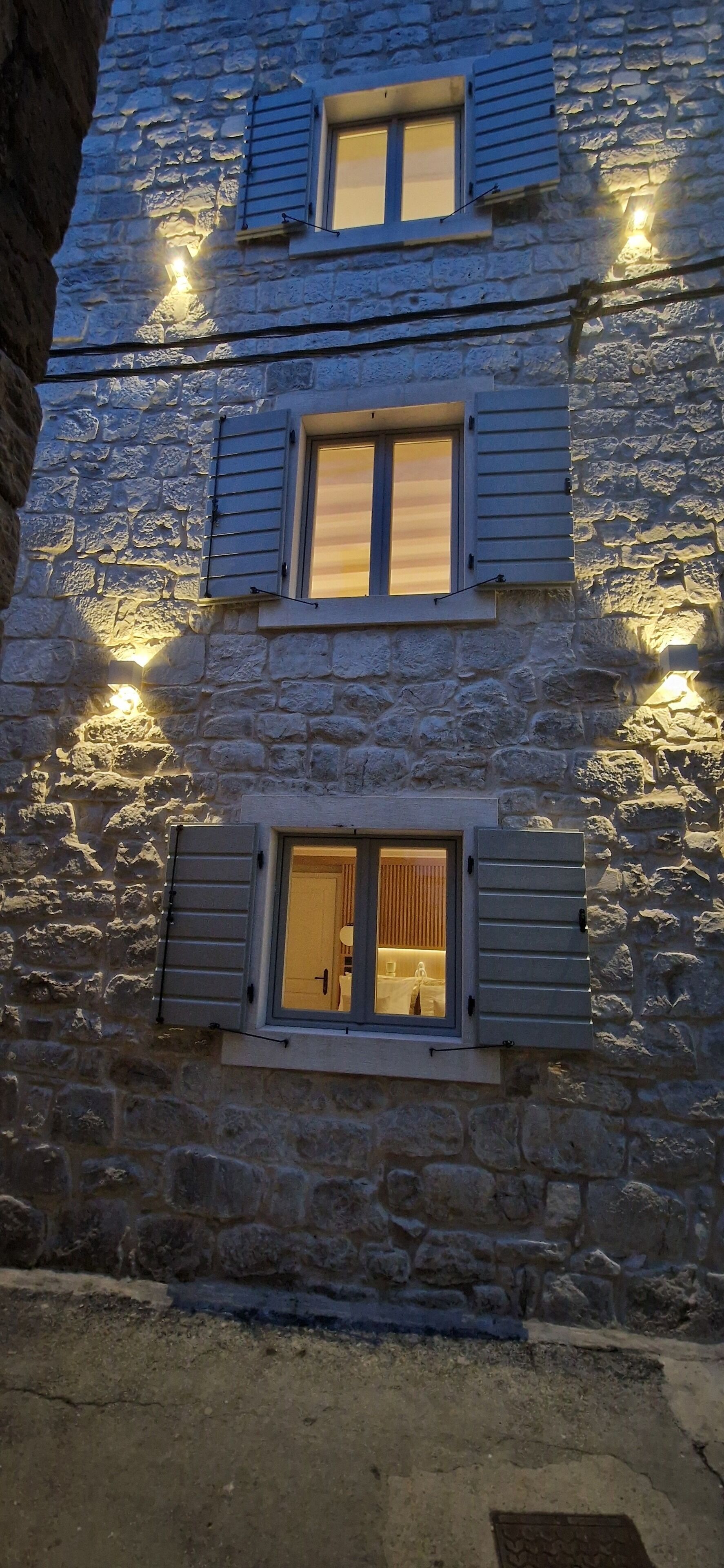 Un edificio de piedra con múltiples ventanas, algunas con contraventanas, e iluminación exterior.