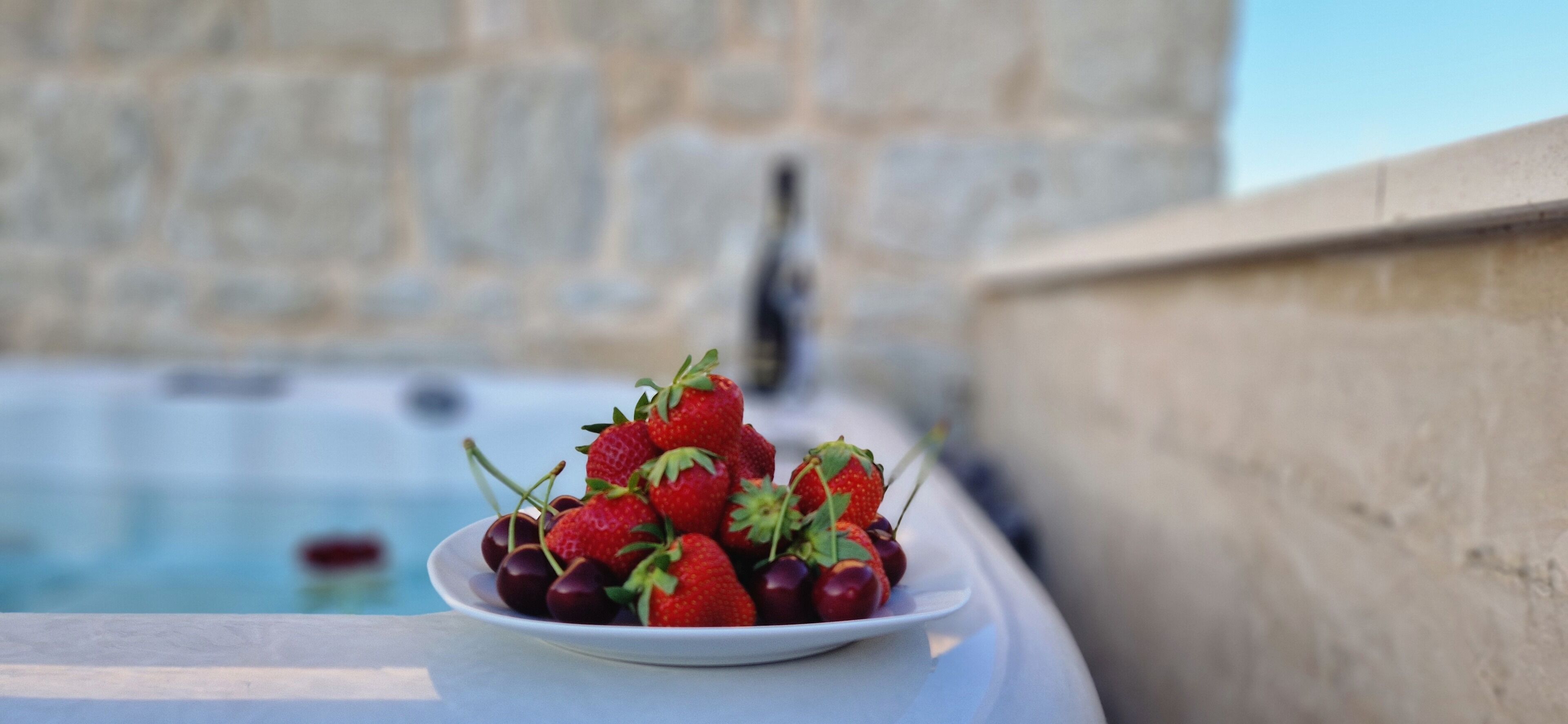 Ein weißer Teller mit Erdbeeren und Kirschen auf einem Tisch in der Nähe einer Steinmauer und eines Pools.