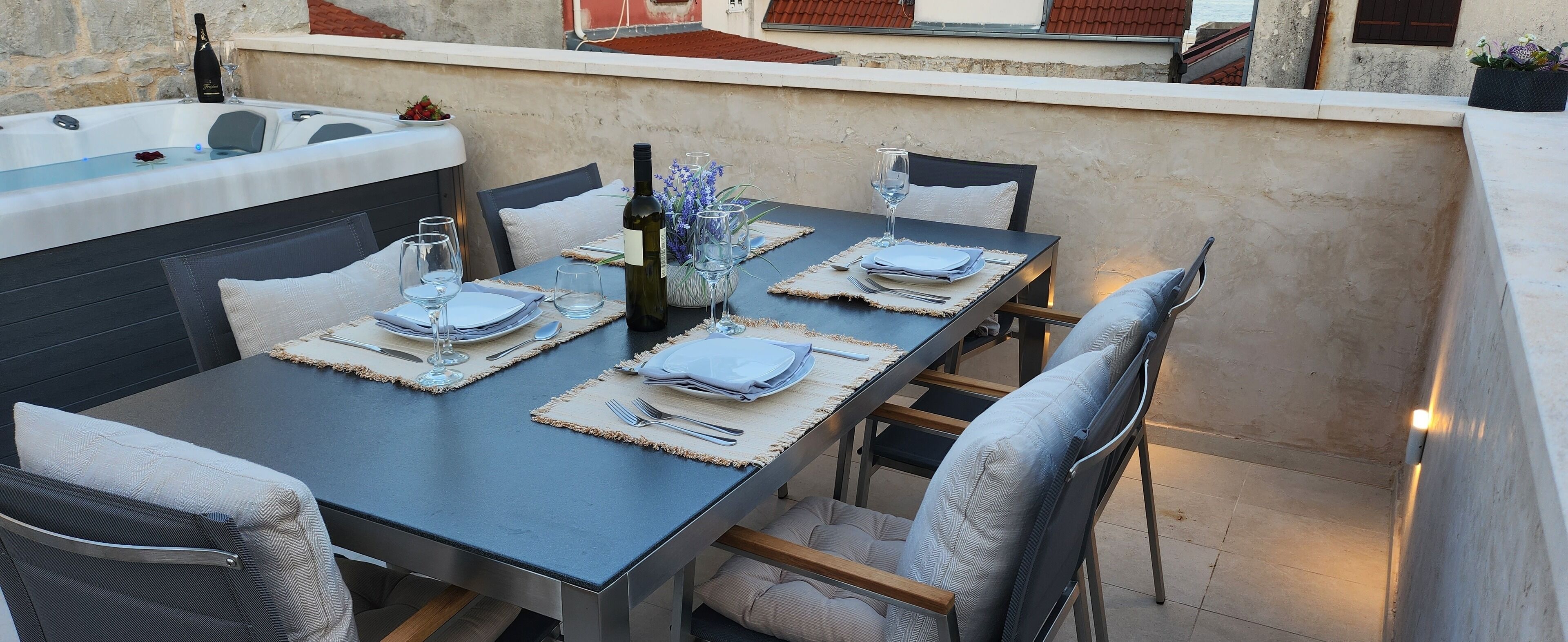 Une table à manger dressée avec des sets de table, des couverts et des verres à vin, avec un jacuzzi et des fleurs en arrière-plan.