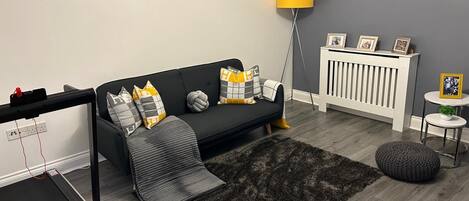 A modern living room with a black sofa, a treadmill, a yellow lamp, and a grey rug.