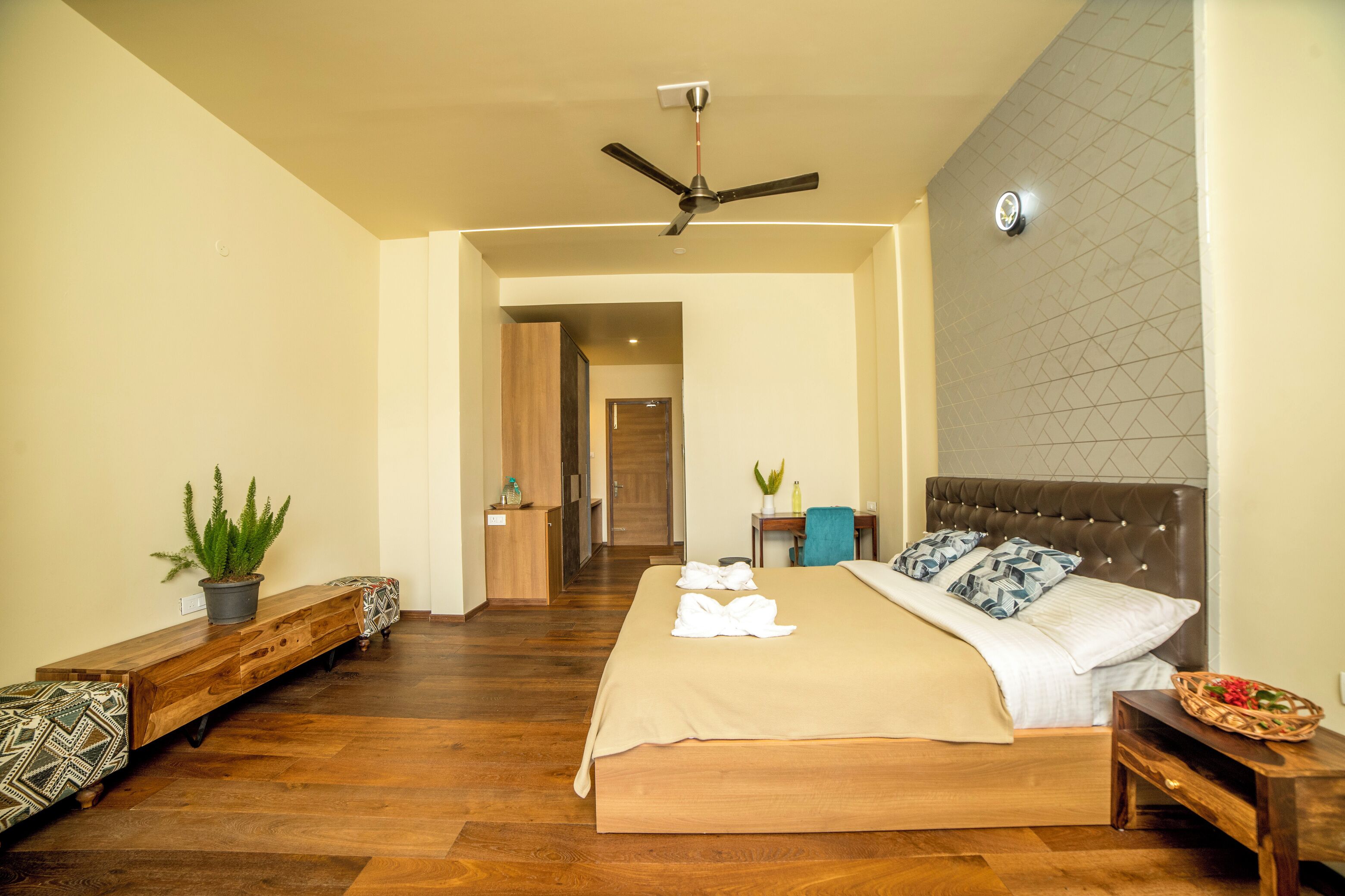 A hotel room with a large bed, a ceiling fan, a wooden bench, and a potted plant.
