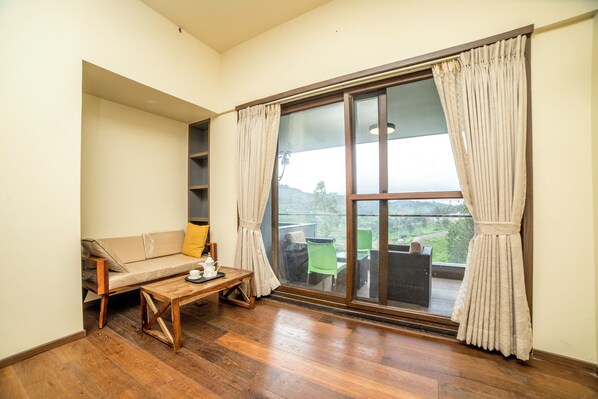 A room with a wooden floor, a sofa, a table with a teapot, and a large window with curtains. - Sri Vidhya Residency (Kotagiri)