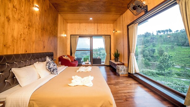 A wooden-paneled bedroom with a large bed, a red sofa, a small table, and a view of a green landscape through a large window. - Sri Vidhya Residency (Kotagiri)