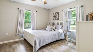Un dormitorio con cama, ventilador de techo, ventana con cortinas, mesita de noche y lámpara.
