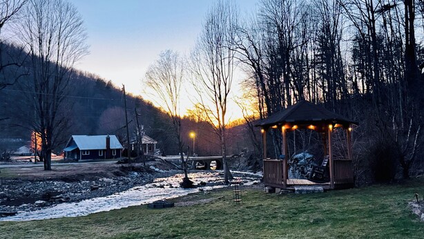 A gazebo in a park with a river, a bridge, and a house in the background. - Charming 1940s Cottage 
Updated-Creek Front-Fire Pit (Bakersville)