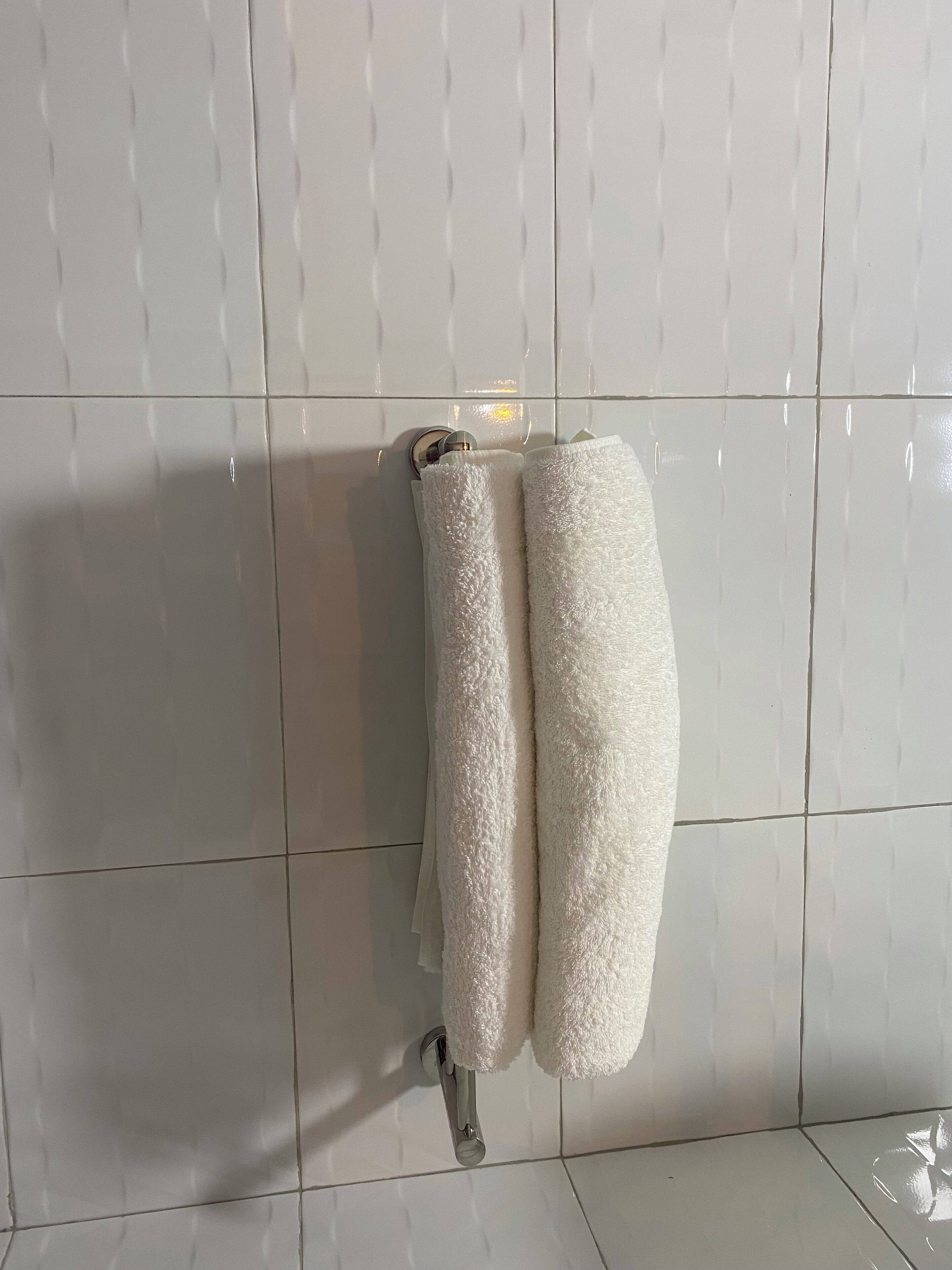 Ein Badezimmer mit weiß gefliesten Wänden und Boden, einem Handtuchhalter mit zwei weißen Handtüchern und einem Chrom-Wasserhahn.