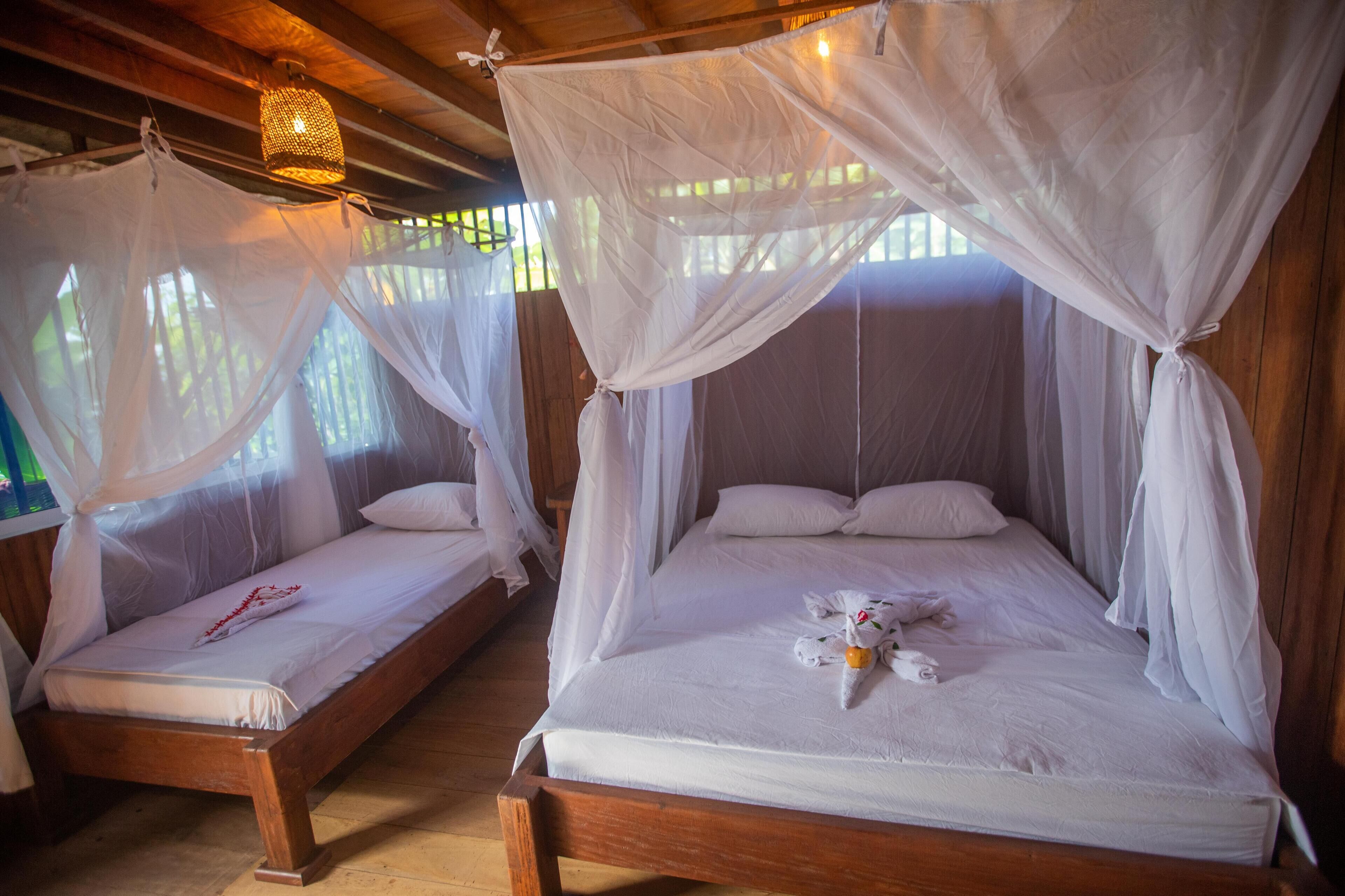 Two wooden beds with white bedding and mosquito netting, one with a red decorative item and the other with a towel and orange fruit.