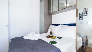 A neatly made bed with a grey blanket, white pillows, a tray with a bottle and glasses, and a small plant. - Piscina academia metro  Brazilian Corner (São Paulo)