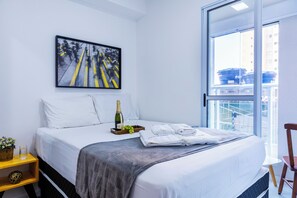 Une chambre d’hôtel avec un lit, une fenêtre, un tableau, une bouteille de champagne, des verres et un plateau de raisins.