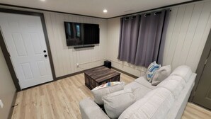 Ein Wohnzimmer mit grauem Sofa, einem Holztisch, einem an der Wand montierten Flachbildfernseher und einer Tür.