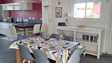 Une cuisine moderne avec des meubles blancs, un coin repas agrémenté d’une nappe à motifs et une bibliothèque remplie de livres.