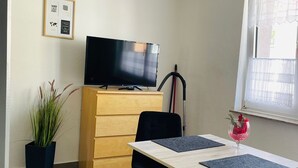 A small, neatly arranged room with a television on a wooden dresser, a potted plant, a dining table with chairs, and a window with sheer curtains. - Quartier 2 - Ferienwohnung Zentral Gelegen (Bremerhaven)
