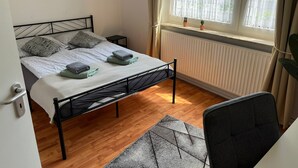 A neatly made bed with pillows and folded towels, a window with a view of greenery, a radiator, and a chair. - Quartier 2 - Ferienwohnung Zentral Gelegen (Bremerhaven)
