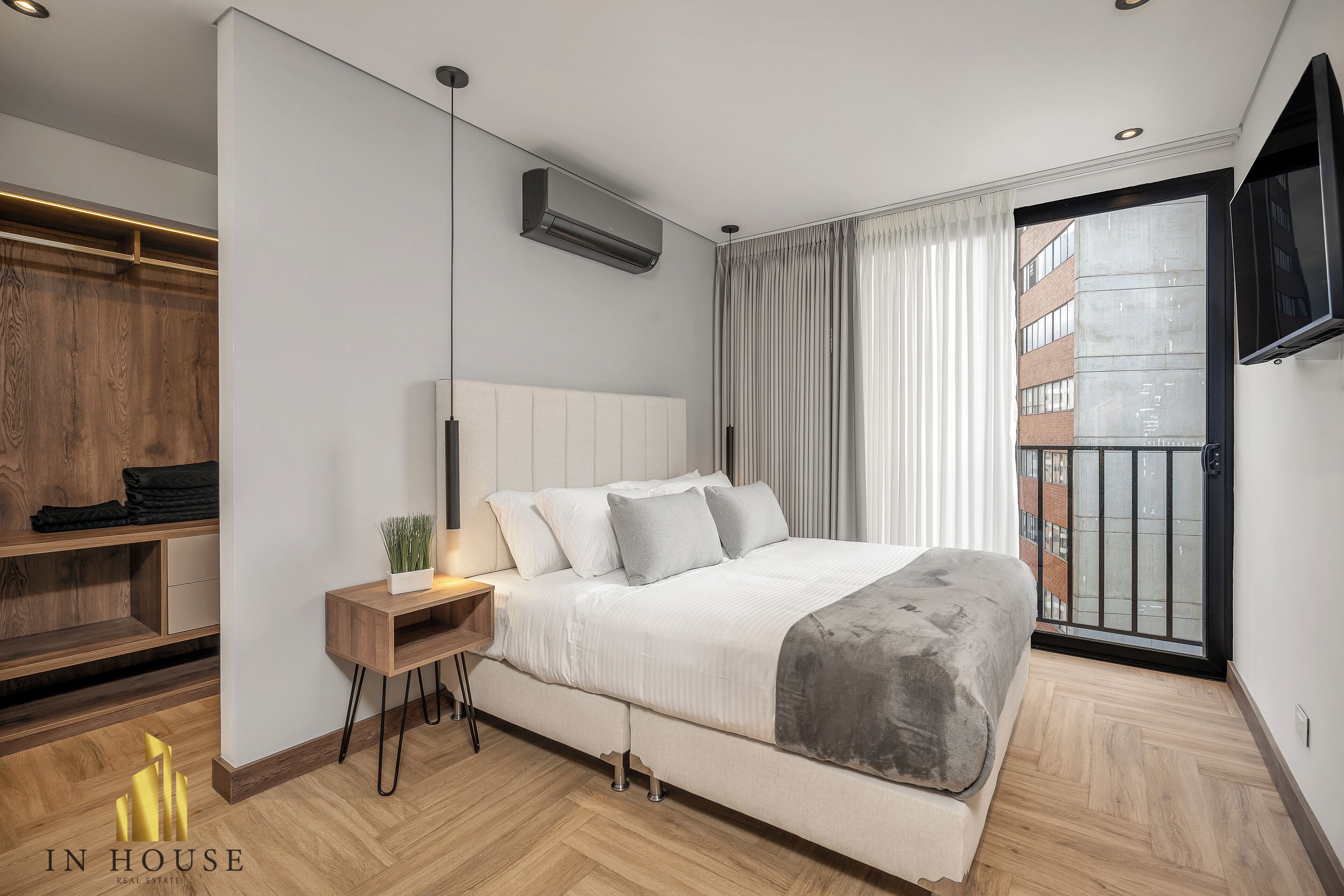 A modern bedroom with a large bed, built-in shelving, a bedside table with a lamp, and a view of the outside through a large window.