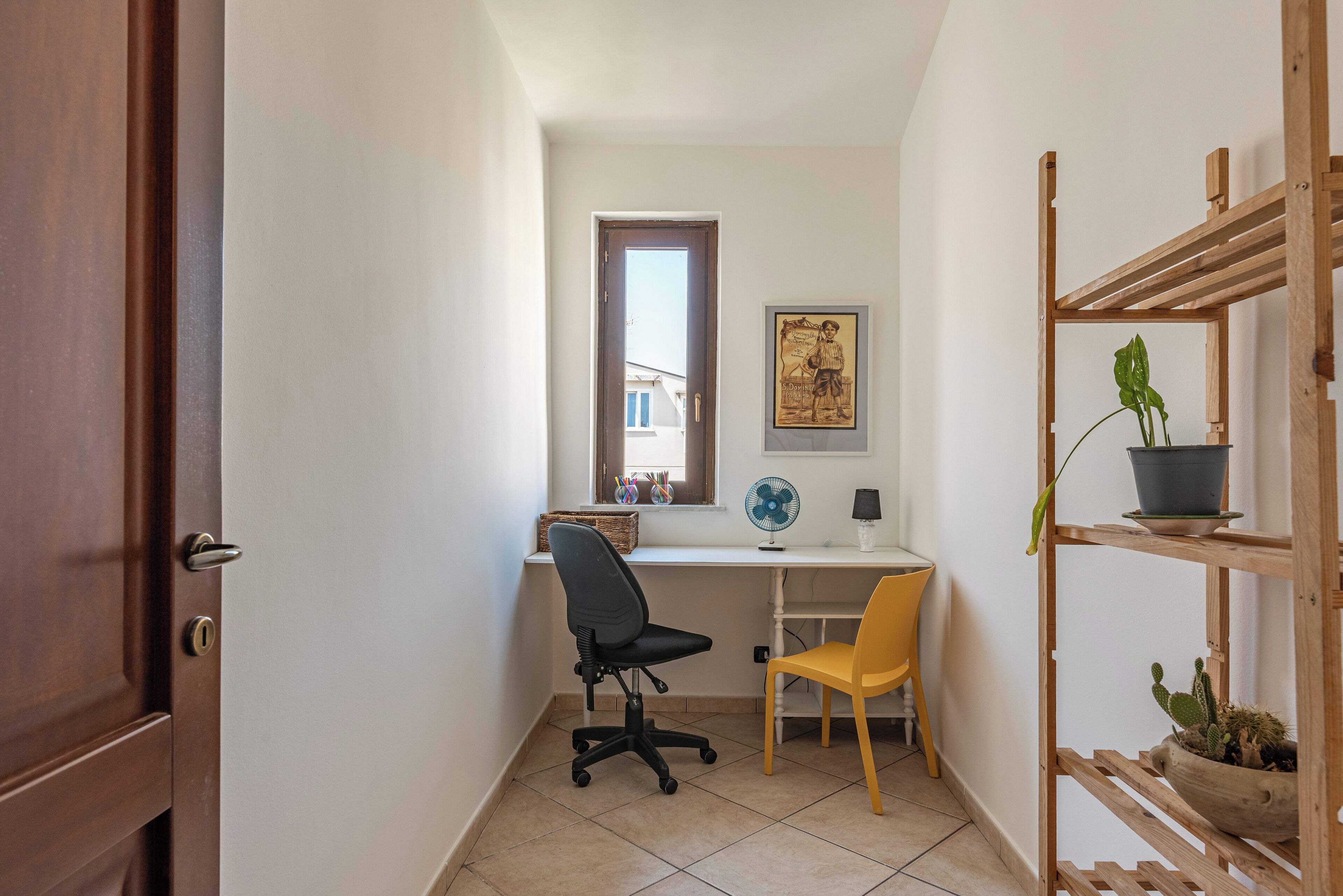 Un petit bureau comprenant un bureau, deux chaises, une fenêtre, un ventilateur et une étagère en bois ornée de plantes.