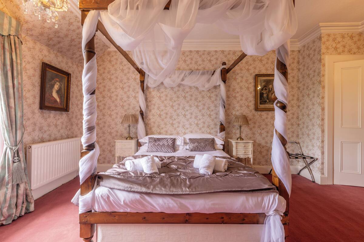 A four-poster canopy bed with a floral wallpaper background, a wooden headboard, and a red carpeted floor.