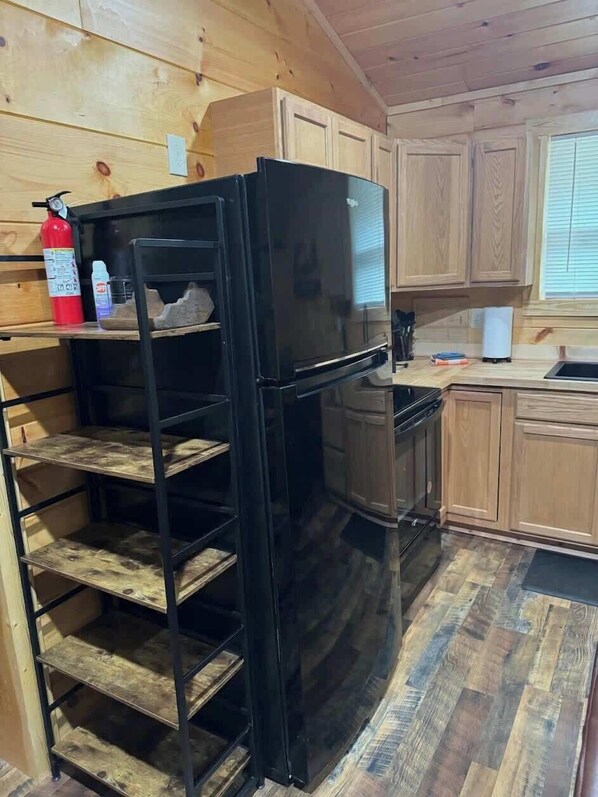 Una cocina compacta con armarios de madera, un refrigerador negro y una estantería metálica.
