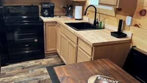 A compact kitchen with wooden cabinets, a black stove, a sink, and a coffee maker. - Cozy Couples Cabin in Heart of RRG! (Campton)