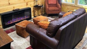 A cozy cabin interior with a leather sofa, a fireplace, a television, and a window with a view of trees. - Cozy Couples Cabin in Heart of RRG! (Campton)