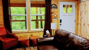 A wooden cabin interior with a leather couch, a red armchair, and a large window offering a view of greenery. - Cozy Couples Cabin in Heart of RRG! (Campton)