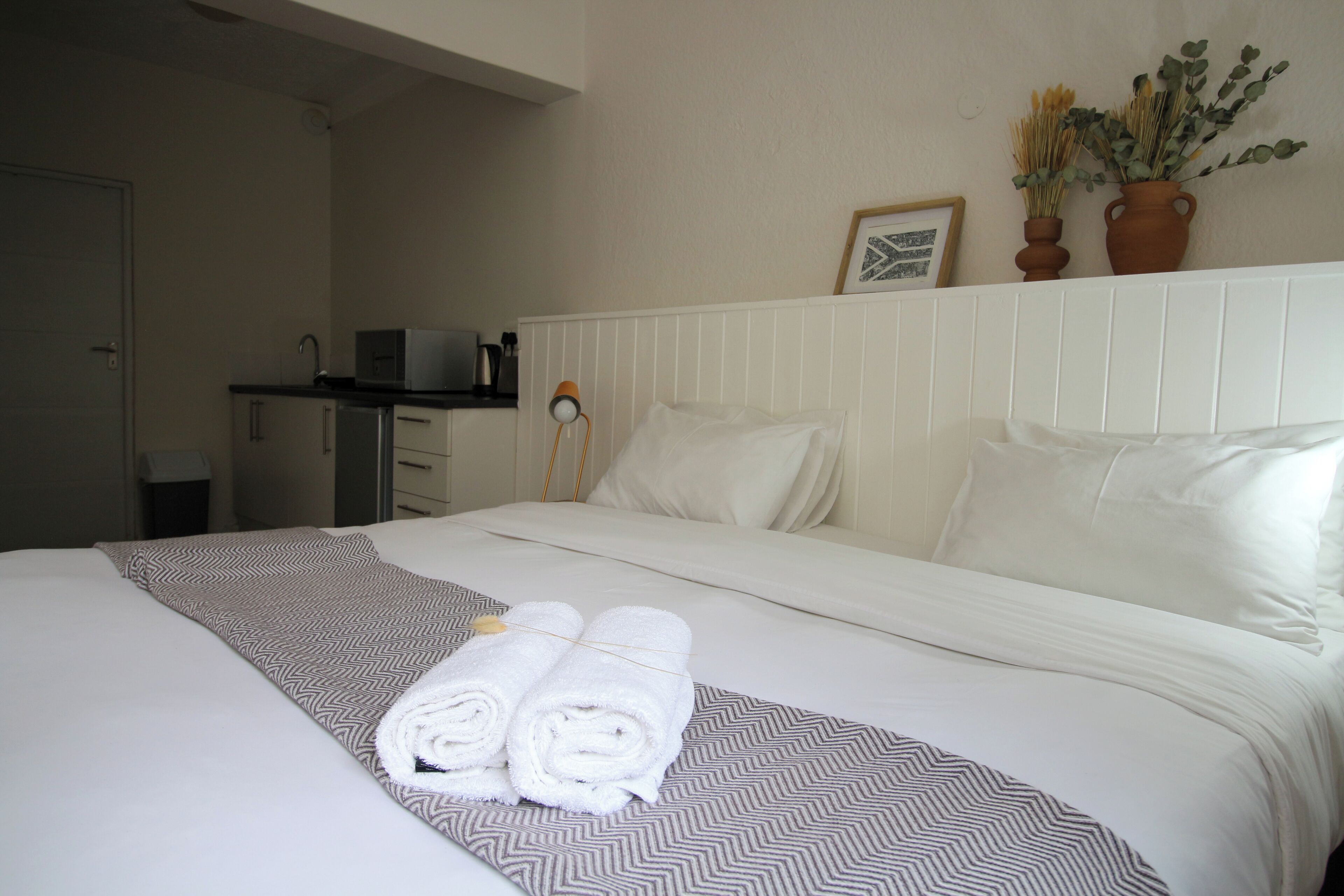 A hotel room with a neatly made bed, a small kitchenette, and a potted plant on the windowsill.