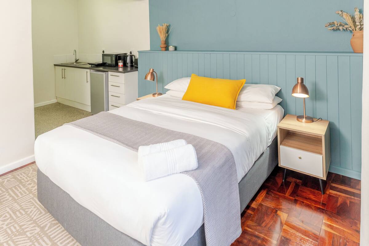 Une chambre comprenant un lit, des tables de chevet, un ventilateur de plafond et une kitchenette située dans la pièce adjacente.