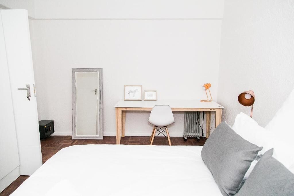 Une chambre à coucher avec un lit, un bureau et une chaise. Il y a un radiateur sous le bureau et une porte sur le côté gauche.