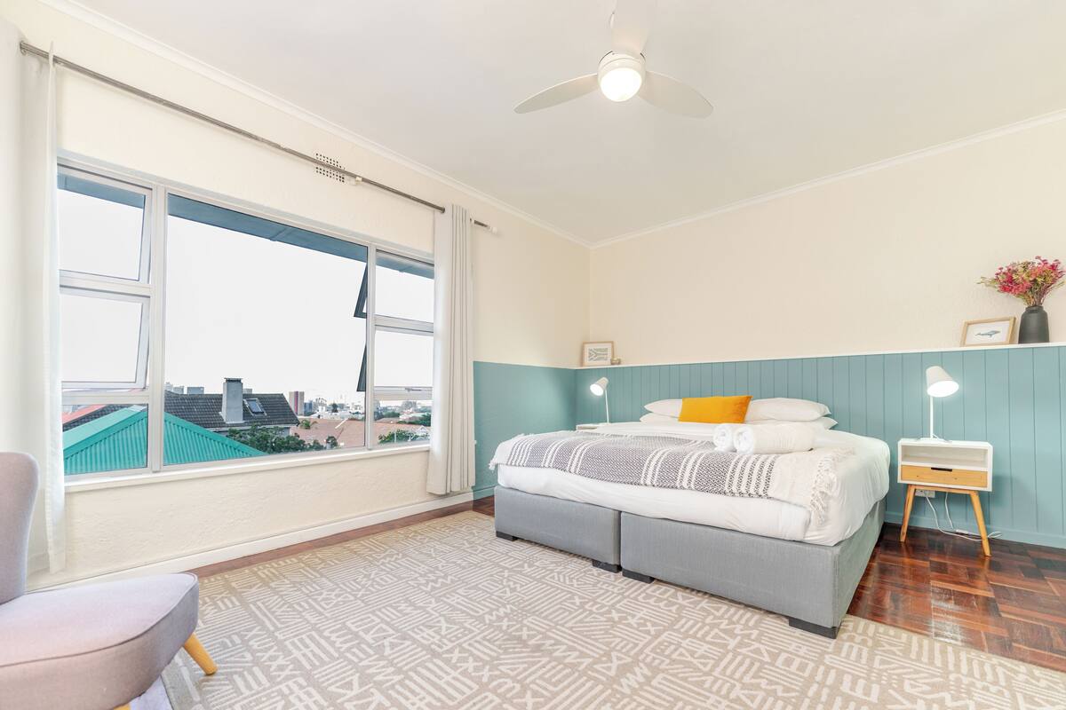 Une chambre à coucher comprenant un lit, une table de chevet, une chaise, une fenêtre avec des rideaux et un ventilateur de plafond.