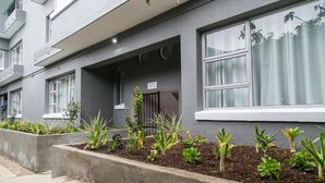 Een modern gebouw met een grijze gevel, grote ramen en een kleine tuin met verschillende planten.