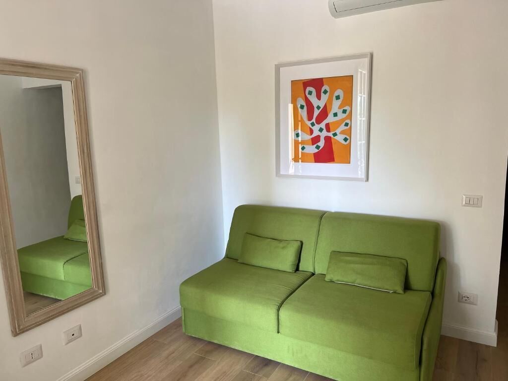A green sofa with two pillows, a framed abstract painting, and a mirror on the wall.