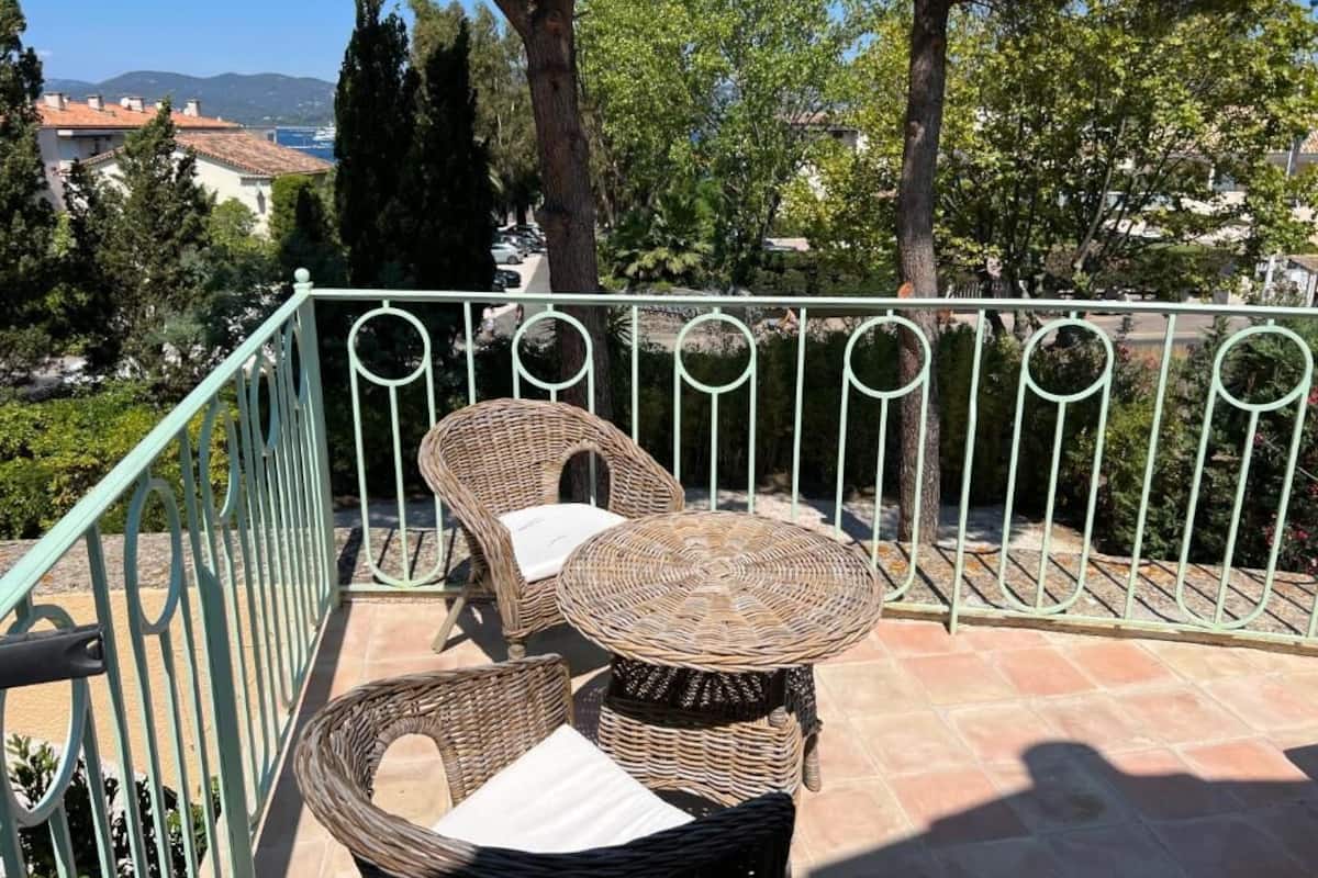 A balcony with wicker furniture, a metal railing, and a view of the sea and buildings in the background.