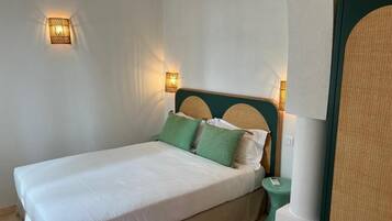 Una cama bien hecha con sábanas blancas y almohadas verdes, una mesita de noche con una lámpara y un aplique de pared.