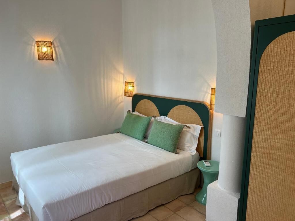 Een net opgemaakt bed met witte lakens en groene kussens, een nachtkastje met een lamp, en een wandlamp.