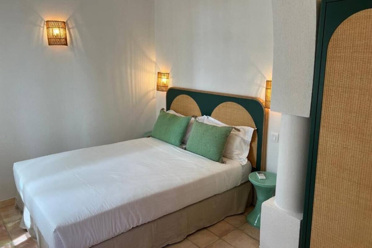 A neatly made bed with white linens and green pillows, a bedside table with a lamp, and a wall-mounted light fixture.