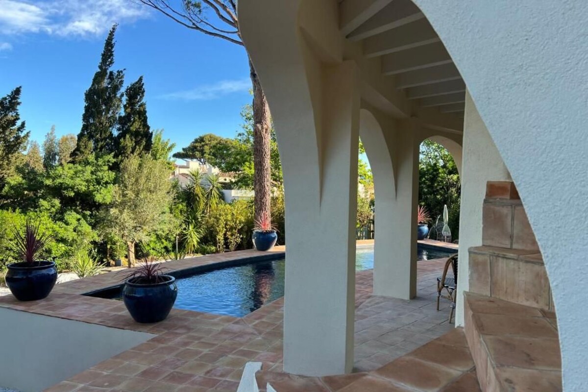 A pool area with a pergola structure, surrounded by potted plants and a view of a residential area.