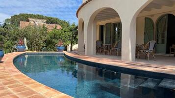 Una piscina de diseño curvo, rodeada por una terraza con baldosas. Hay sillas al aire libre y una zona de estar techada con barandilla.