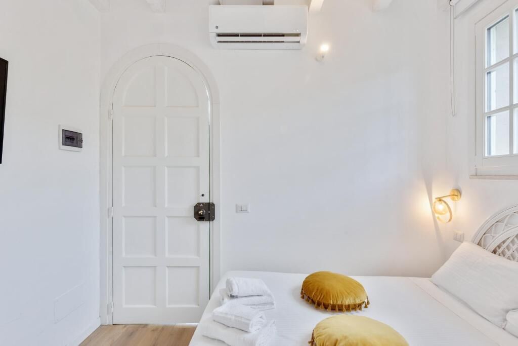 Une chambre avec une porte blanche, un lit recouvert de draps blancs, un oreiller jaune et un luminaire fixé au mur.