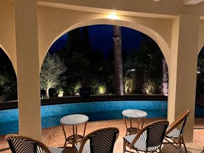 Zona junto a la piscina con vista a árboles y cielo nocturno. Hay sillas y mesas bajo un arco.