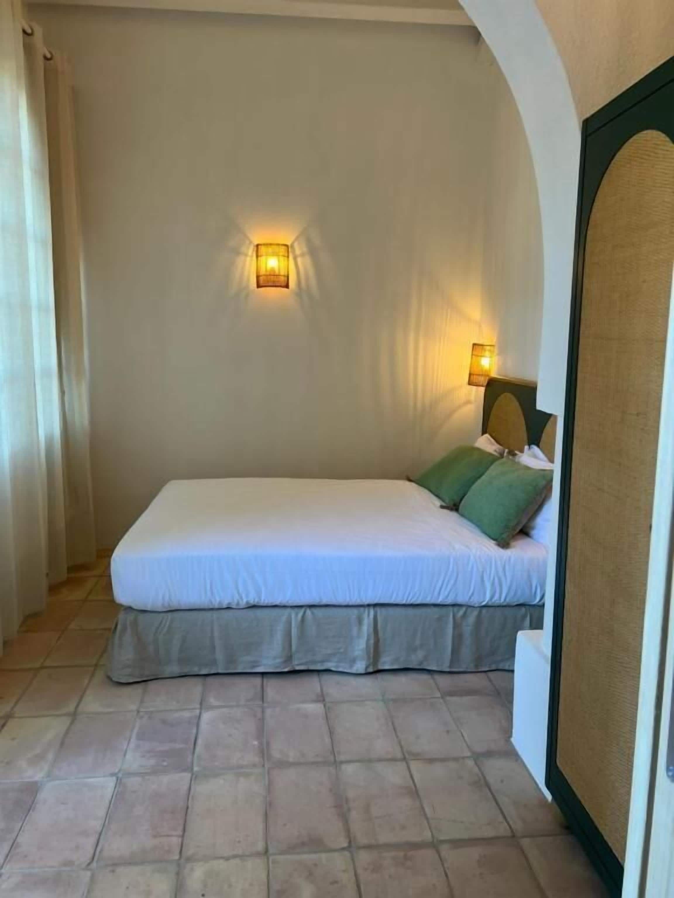 A single bed with a white mattress and green pillows, a wall-mounted light, and a tiled floor.