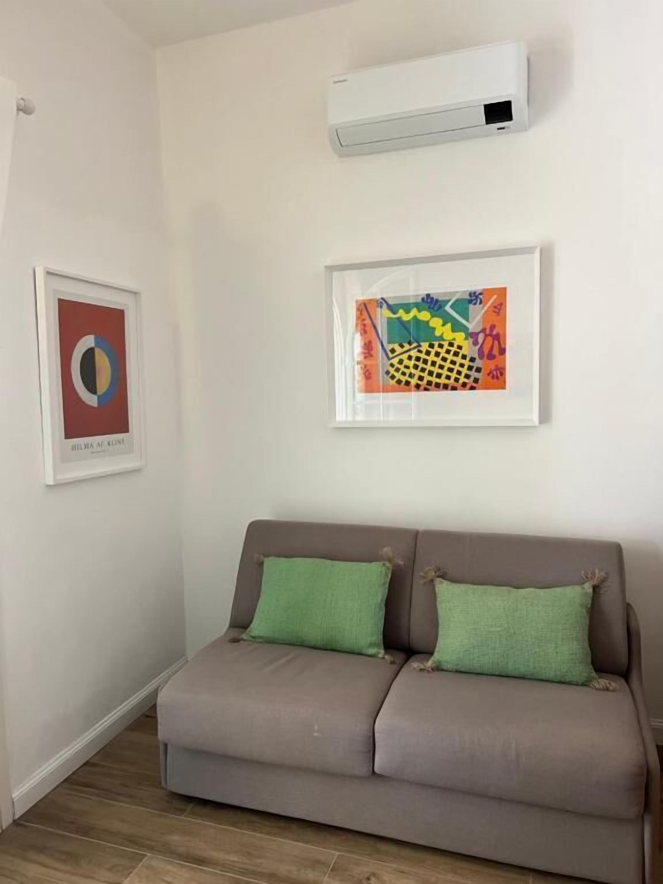 Uma sala de estar com um sofá cinza, duas almofadas verdes, um aparelho de ar condicionado branco e duas obras de arte emolduradas na parede.
